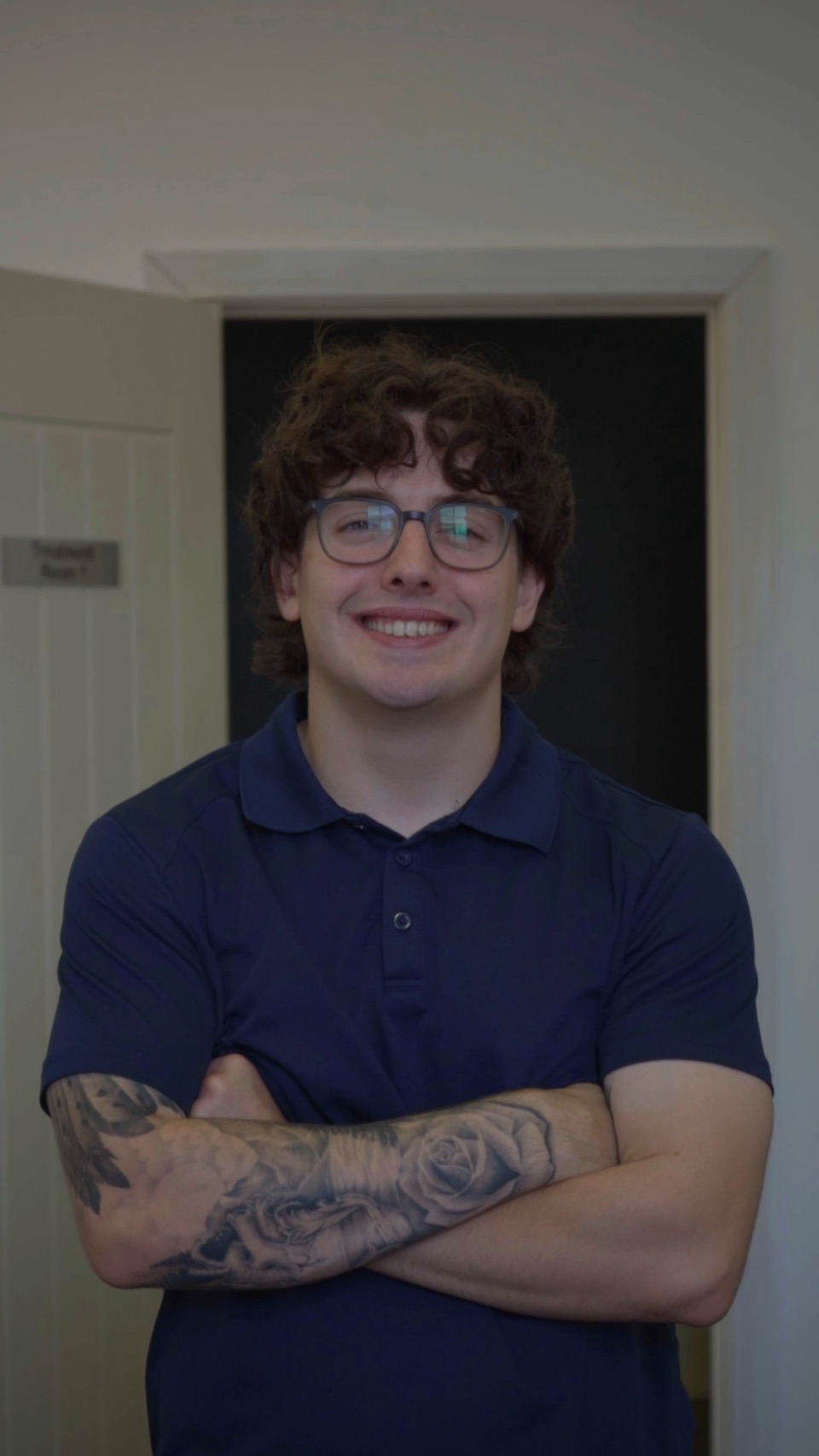 A chiropractor in a navy polo shirt, arms folded, smiling at the camera from a doorway at the clinic